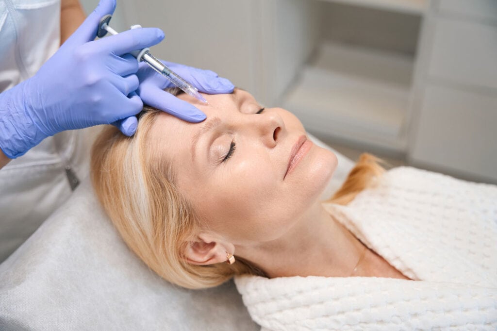 Close up of face clinic speclialist hands performing rejuvenating injection on mature lady forehead in botox clini